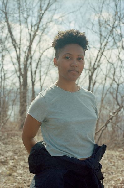 Photo by the poet, titled Derica Climbing Beacon, which in the foreground features a portrait of a Black woman with short hair and in the background features bare trees that have shed brown leaves onto the ground